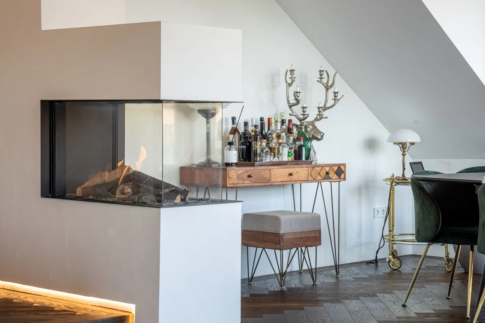 Wohnzimmer mit modernem Kamin, Barwagen, Regalen mit Flaschen und Hockern auf Holzfußboden.