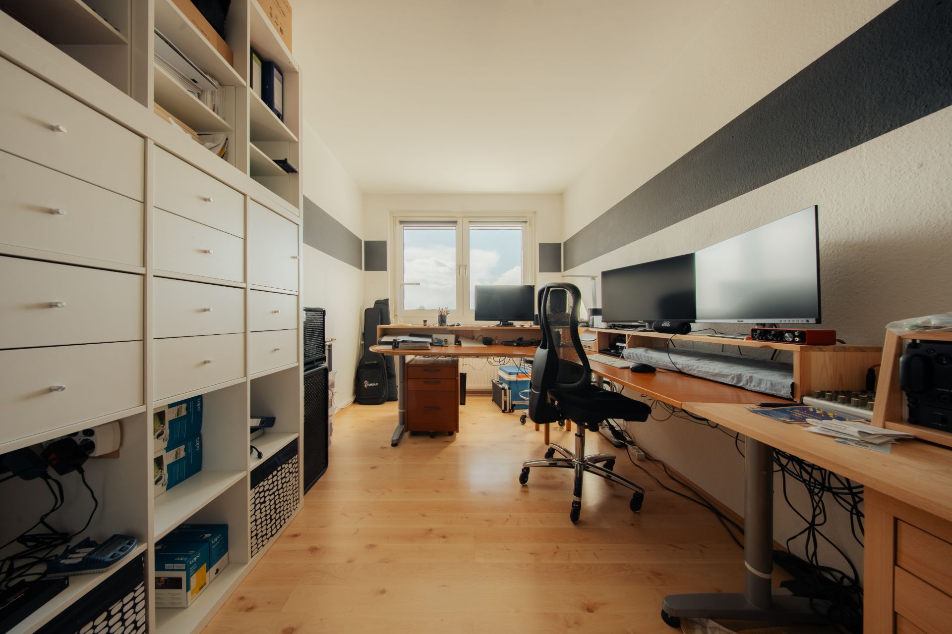 Arbeitszimmer mit Regalen, Schreibtischen, Stühlen und mehreren Monitoren, großes Fenster im Hintergrund.