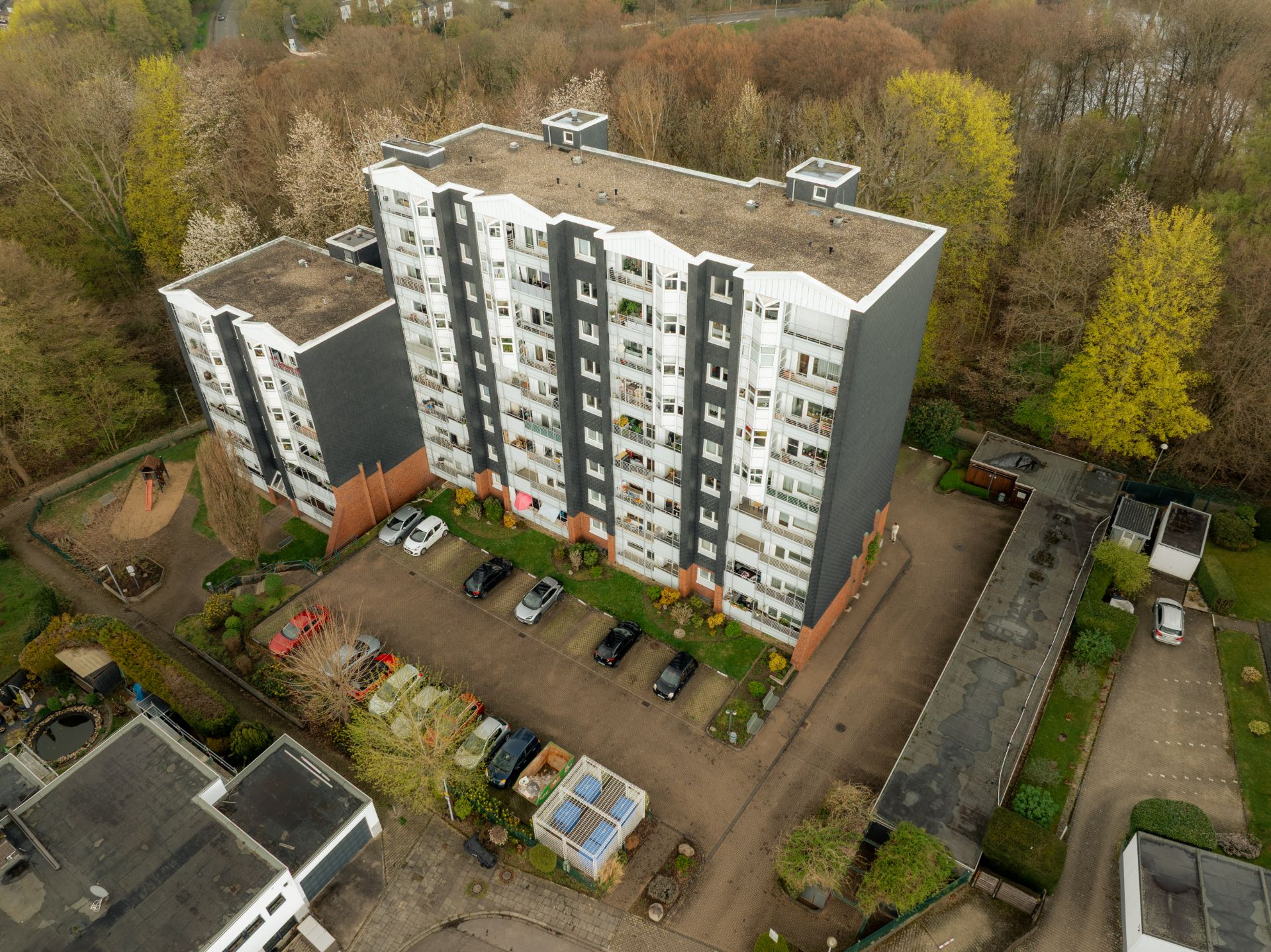 Mehrstöckiges Wohngebäude mit Parkplatz, umgeben von Bäumen und anderen Gebäuden.