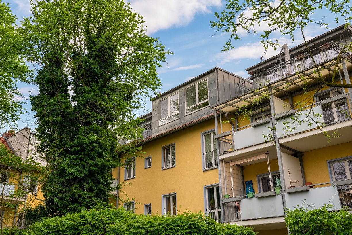 Gelbes Wohnhaus mit Balkonen und großen Fenstern, umgeben von Bäumen und Sträuchern.