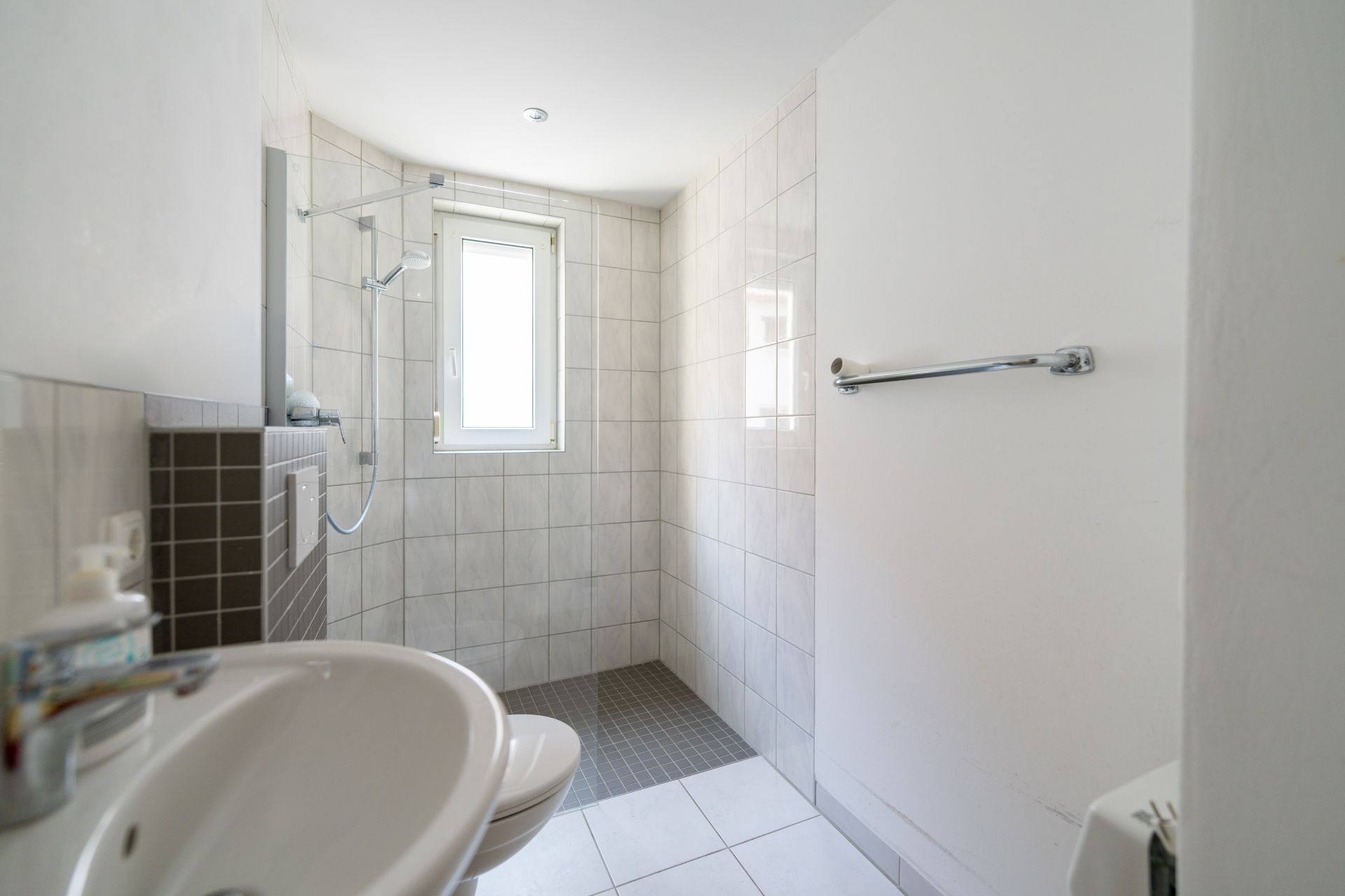 Helles, gefliestes Badezimmer mit bodengleicher Dusche, Waschbecken und Fenster.
