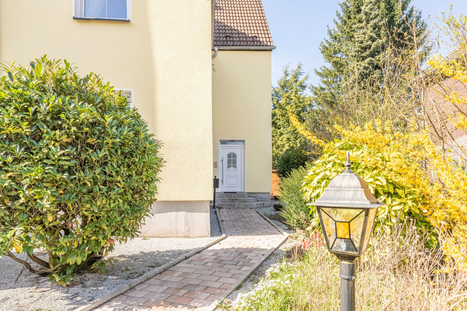 Gepflasterter Weg führt zu einer weißen Haustür, umgeben von Sträuchern und Gartenlaterne.