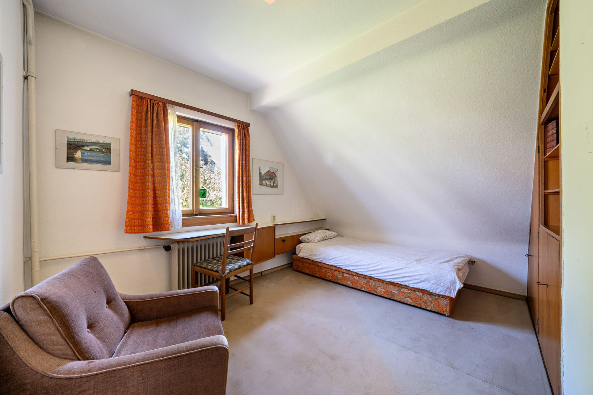 Helles Zimmer mit Einzelbett, braunem Sessel, Holzstuhl und Fenster mit orangefarbenen Vorhängen.