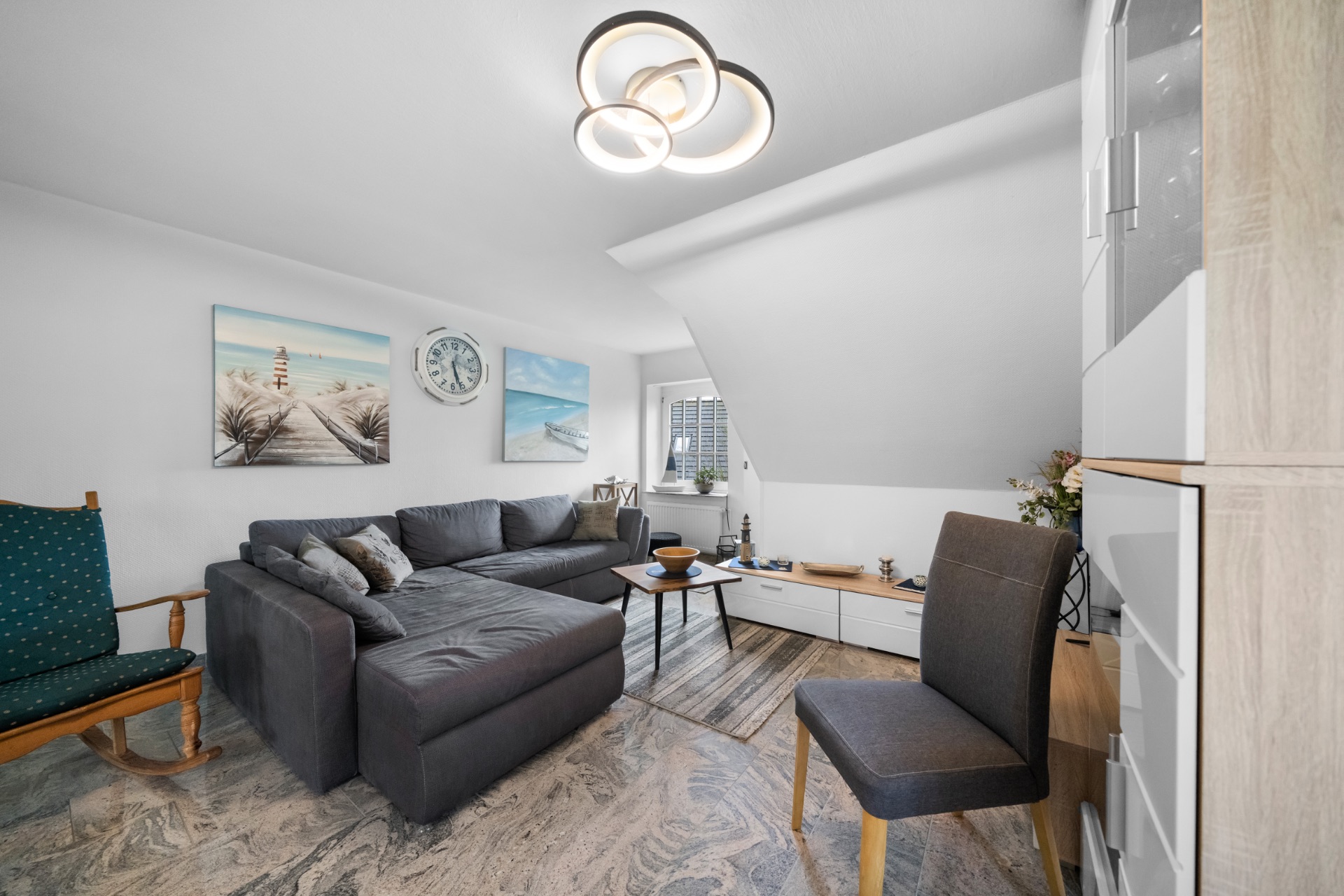 Helles Wohnzimmer mit grauem Ecksofa, Schaukelstuhl, Wandbildern und modernem Kronleuchter.