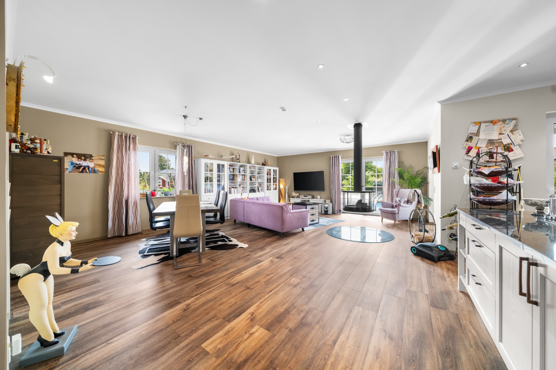 Großes Wohnzimmer mit Holzfußboden, Sofa, Esstisch, Bücherregal und großen Fenstern.