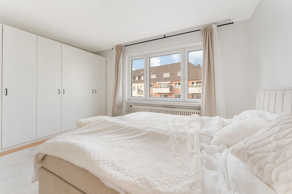 Helles Schlafzimmer mit großem Bett, weißen Schränken und Fenster mit Blick auf Nachbarhäuser.