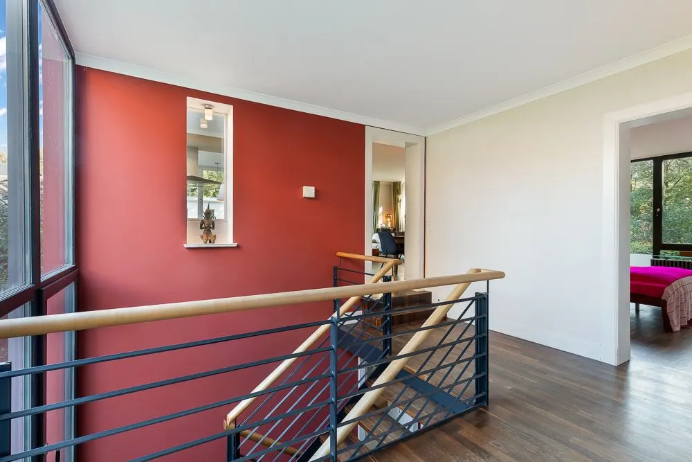 Heller Flur mit Treppe, roter Wand und offenem Zugang zu einem Zimmer mit Bett.