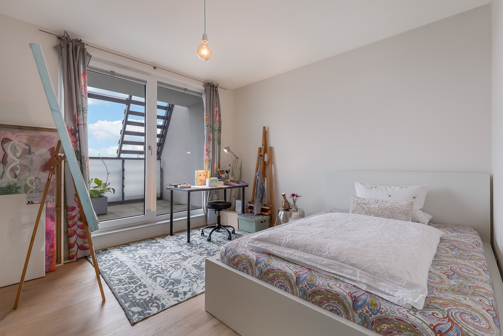 Helles Schlafzimmer mit Bett, Schreibtisch am Fenster, Staffelei und Balkonzugang.