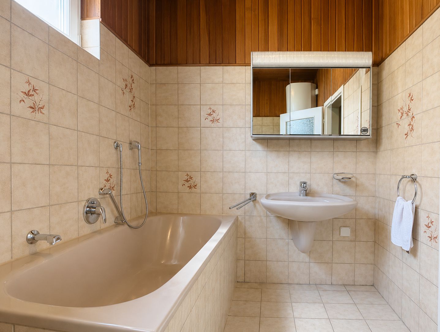 Badezimmer mit Badewanne, Waschbecken, Spiegel und beigefarbenen Fliesen mit Blumenmuster.