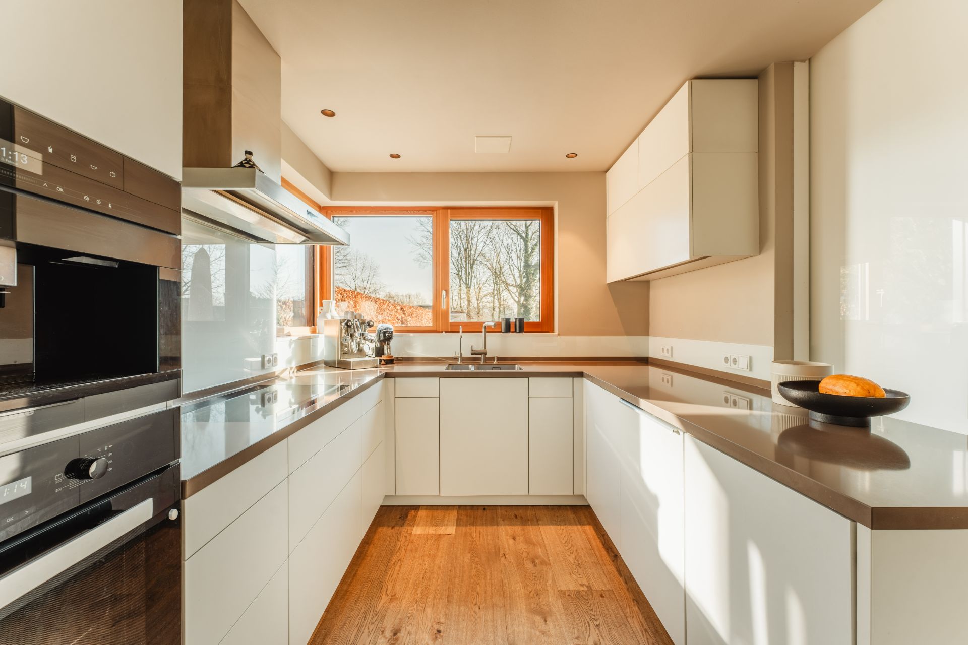 Moderne, helle Einbauküche mit großen Fenstern, weißen Schränken und Holzfußboden.
