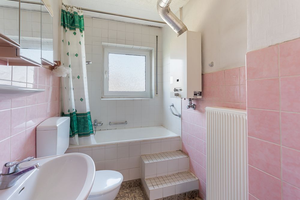 Kleines Badezimmer mit Badewanne, Waschbecken, Toilette, rosa Fliesen und Fenster.