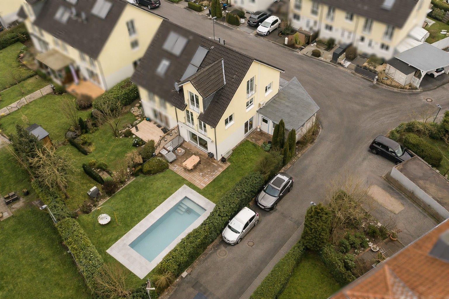Luftaufnahme eines gelben Wohnhauses mit Garten, Terrasse, Pool und zwei geparkten Autos auf der Straße.