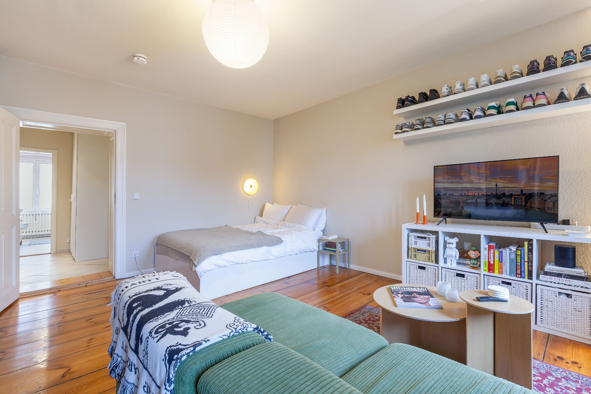 Helles Schlafzimmer mit Doppelbett, Sofa, Fernseher, Bücherregal und Schuhregalen an der Wand.