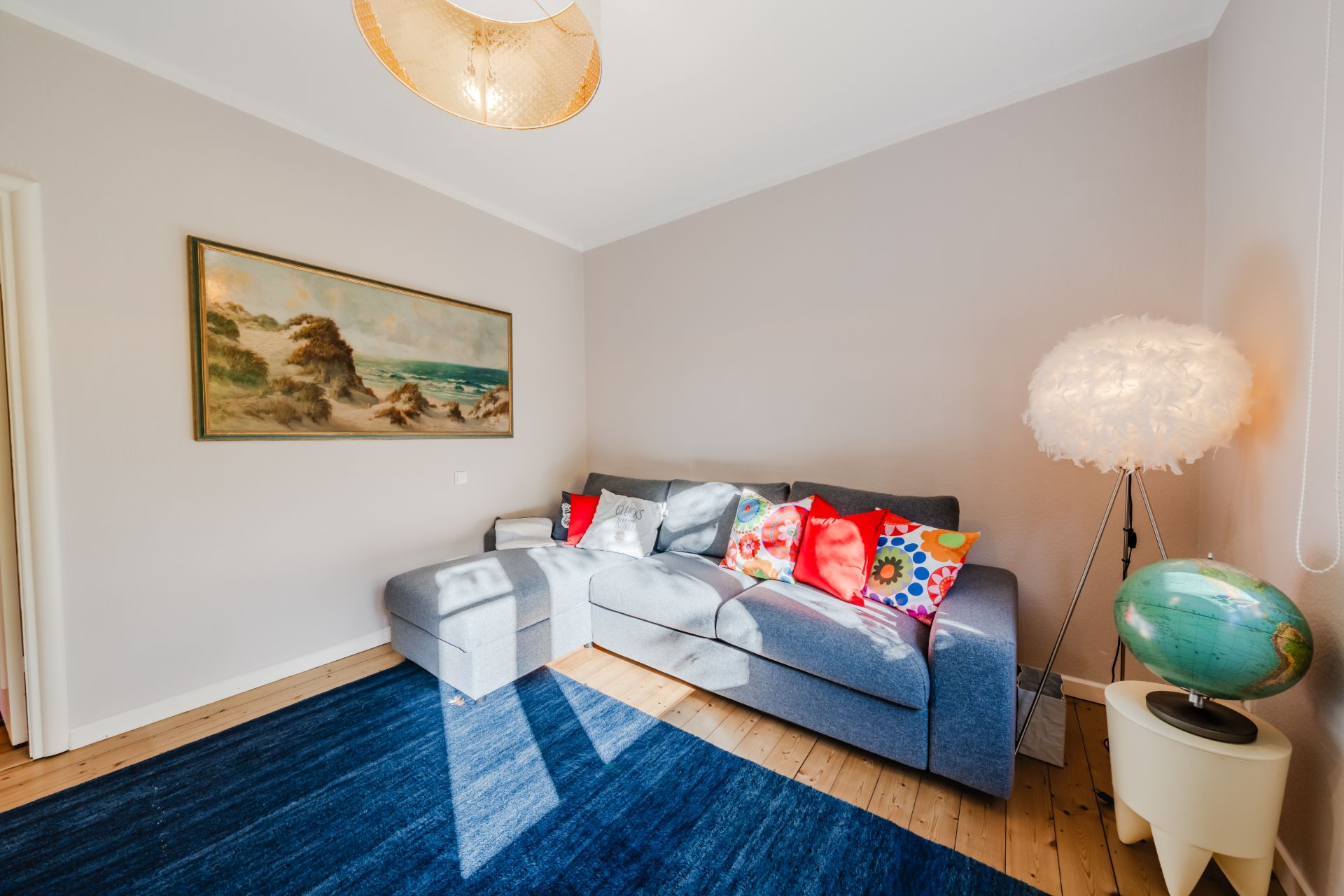 Helles Wohnzimmer mit blauem Teppich, grauem Sofa, bunter Kissen und Stehlampe mit Feder-Lampenschirm.