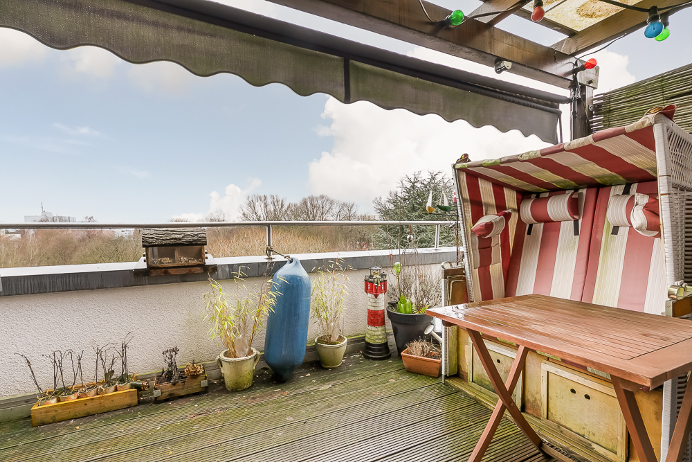 Balkon mit Sonnenschutz, Strandkorb, Holztisch, Pflanzentöpfen und Blick ins Grüne.