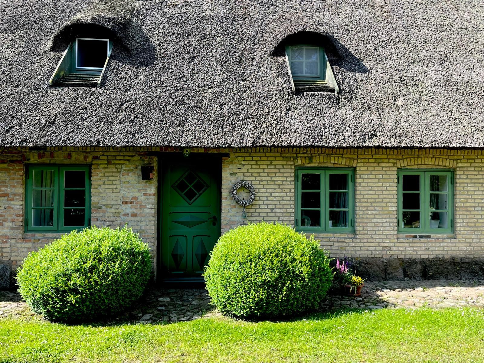 Backsteinhaus mit Reetdach, grüner Tür und Fenstern, zwei Büschen im Vorgarten.