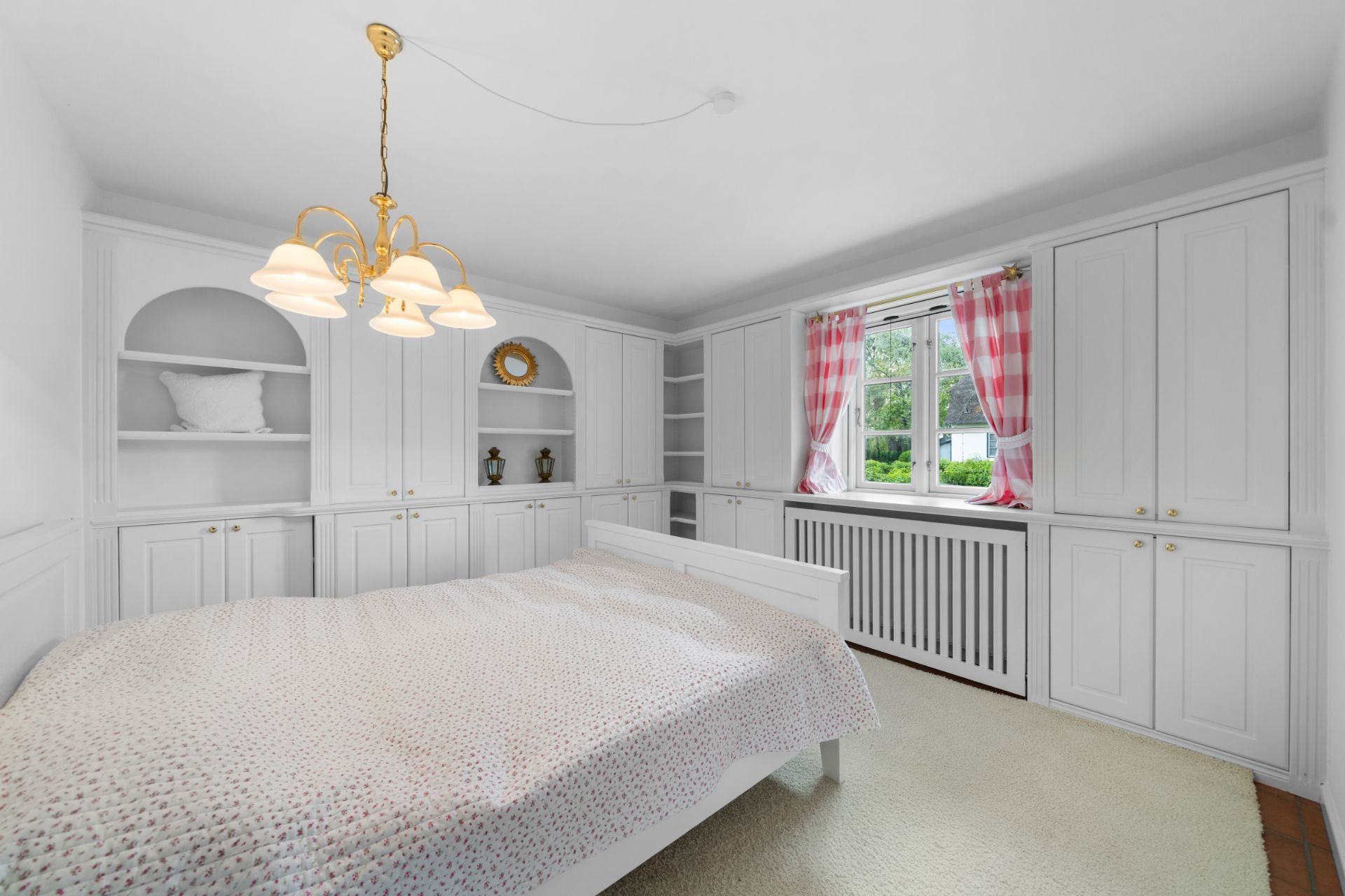 Helles Schlafzimmer mit weißem Bett, Einbauschränken und Fenster mit roten Vorhängen.