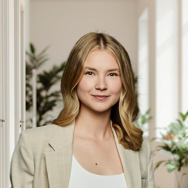 Junge Frau in beigem Anzug steht in einem hellen Raum mit Pflanzen am Fenster.