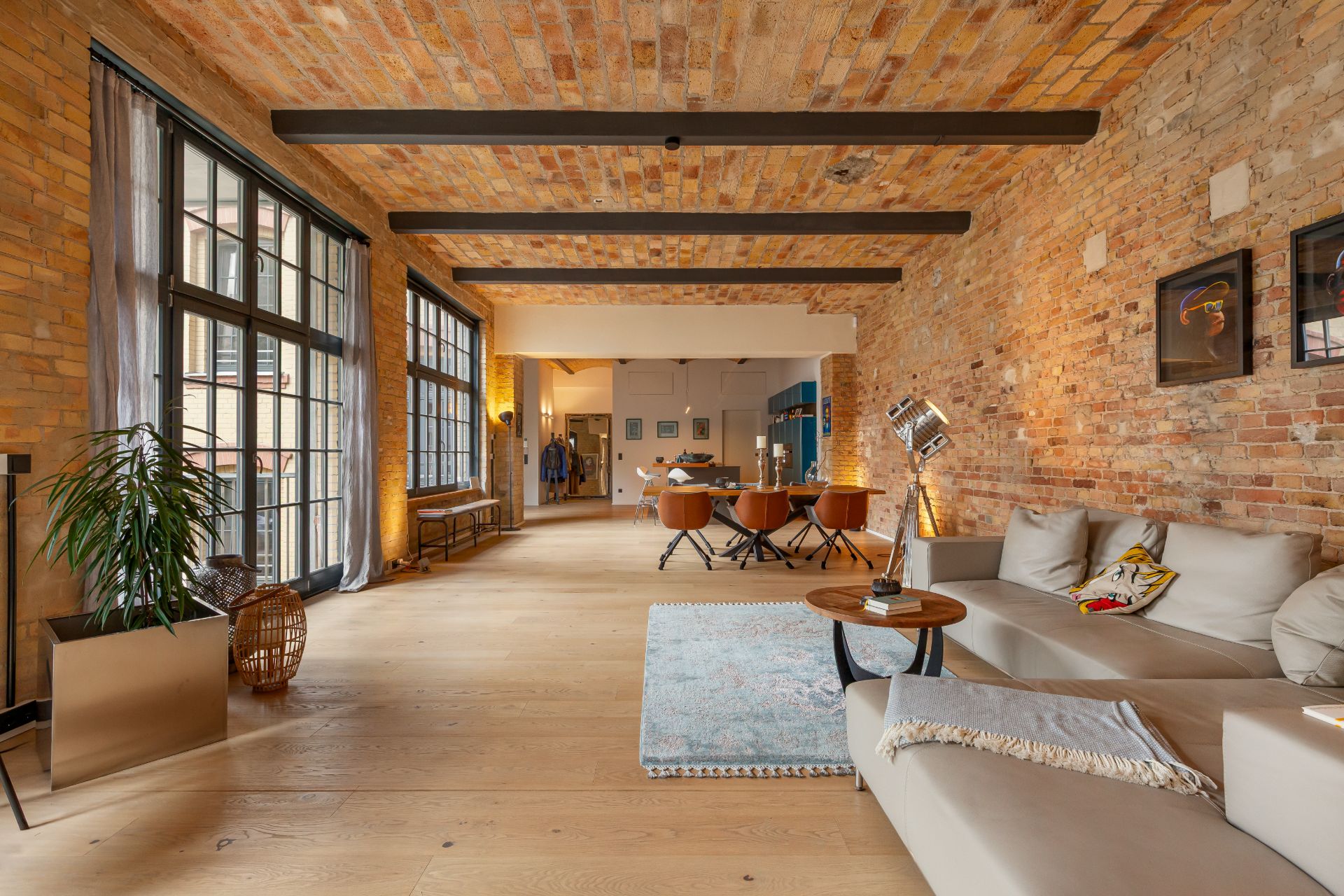 Helles Wohnzimmer mit Ziegelwänden, großen Fenstern, Sofa, Teppich und Esstisch mit Stühlen.