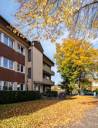 Mehrfamilienhaus mit Balkonen, Herbstbäumen und parkenden Autos auf einer von Laub bedeckten Straße.