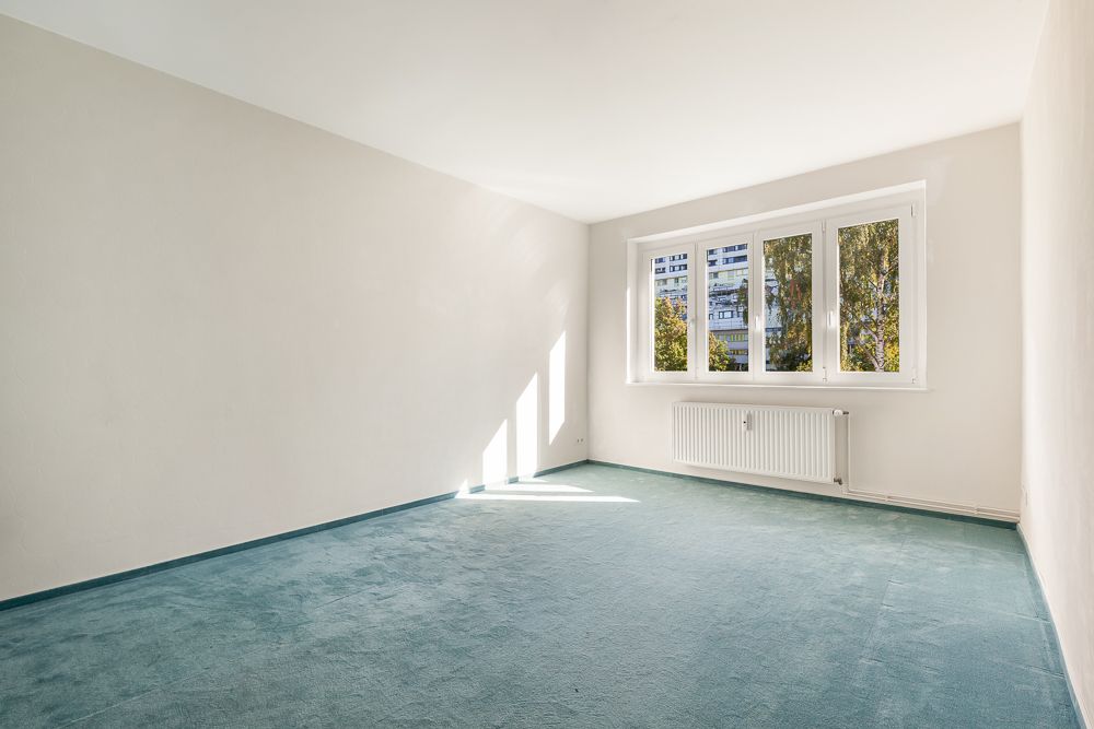 Helles, leeres Zimmer mit blauem Teppich und großem Fenster mit Blick nach draußen.