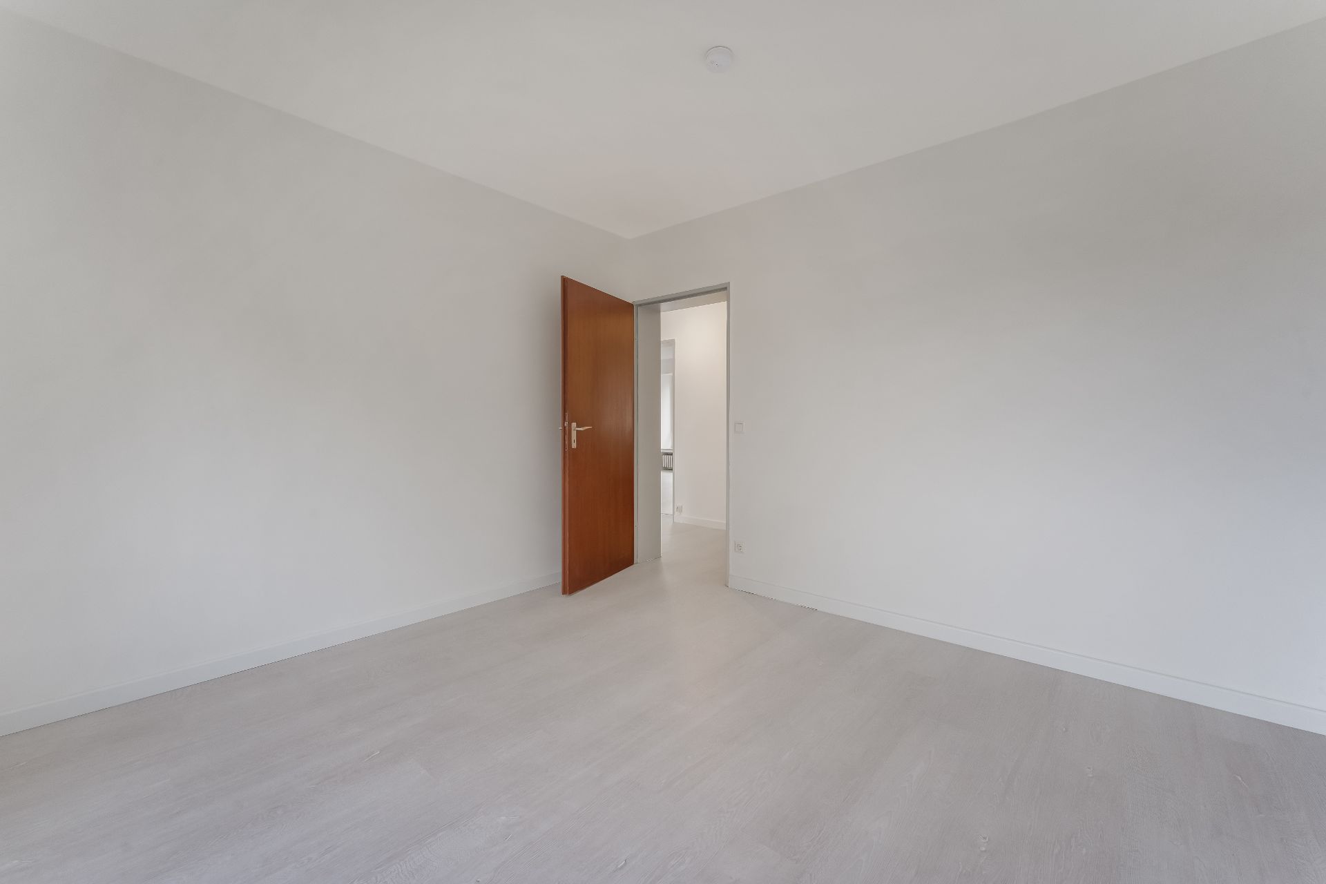 Leerer Raum mit weißem Boden und weißen Wänden, offene Holztür führt in einen Flur.