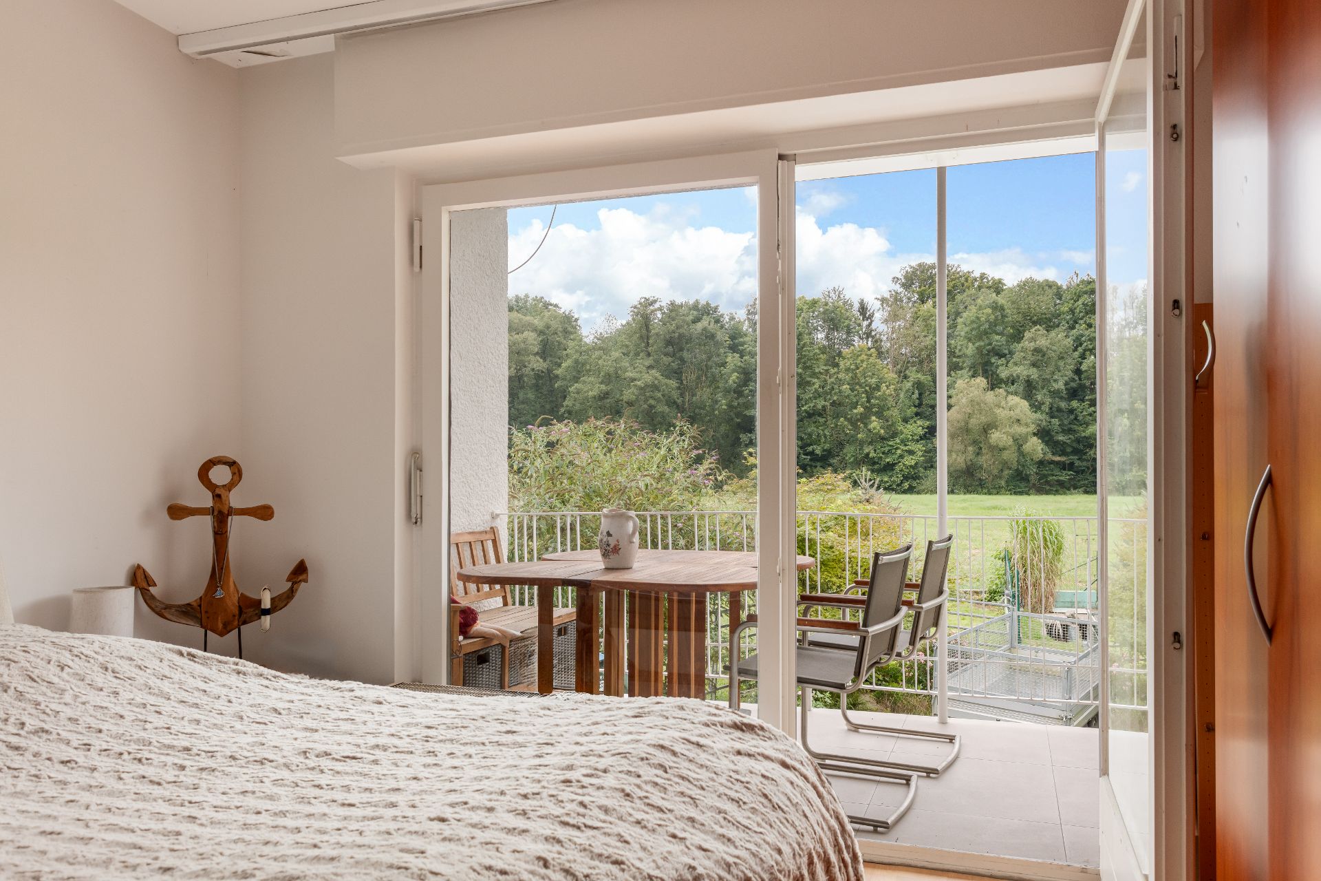Schlafzimmer mit Bett, Holzschrank und Zugang zum Balkon mit Tisch und Stühlen, Blick ins Grüne.