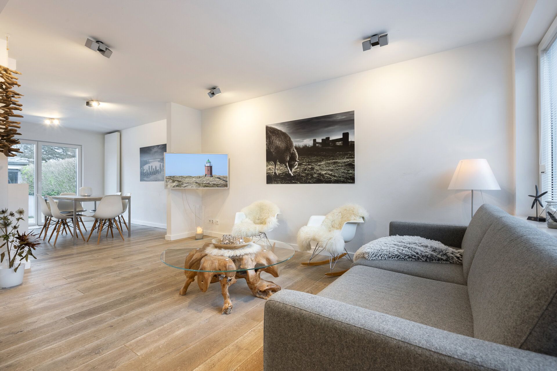 Modernes Wohnzimmer mit grauem Sofa, Glastisch, Holzboden und Essbereich im Hintergrund.