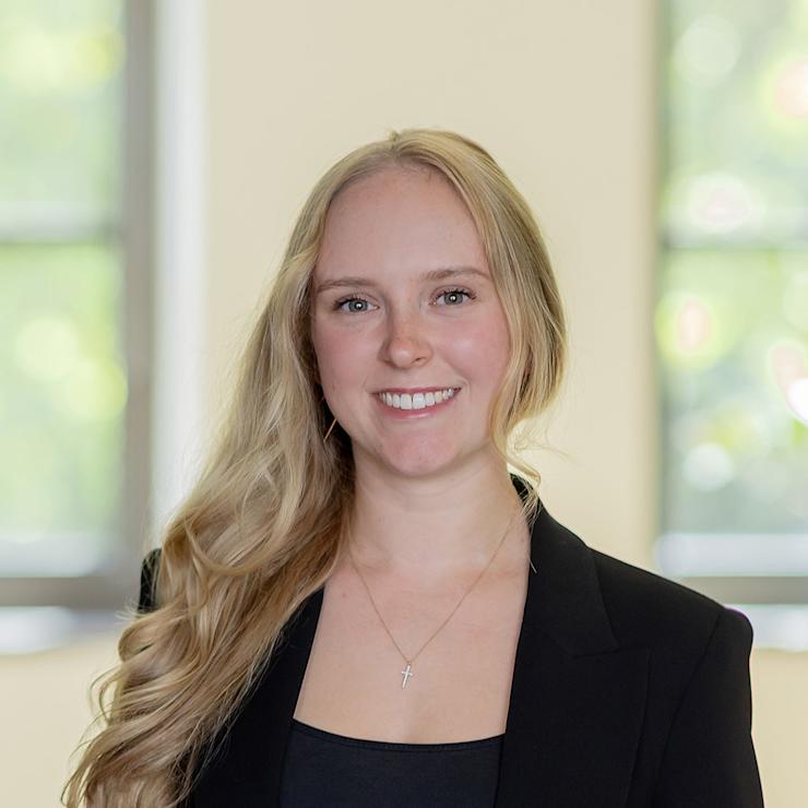 Frau mit langen blonden Haaren und schwarzer Kleidung steht vor einem unscharfen Fensterhintergrund.