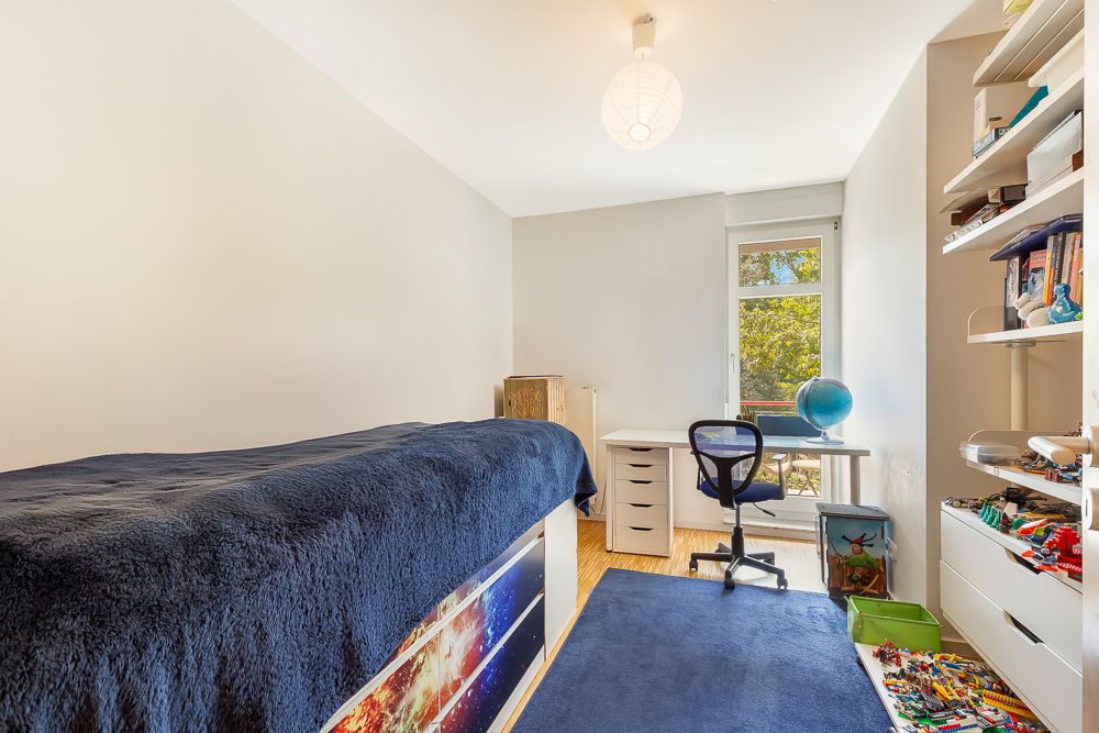 Helles Kinderzimmer mit Bett, Schreibtisch, Regal und großem Fenster.