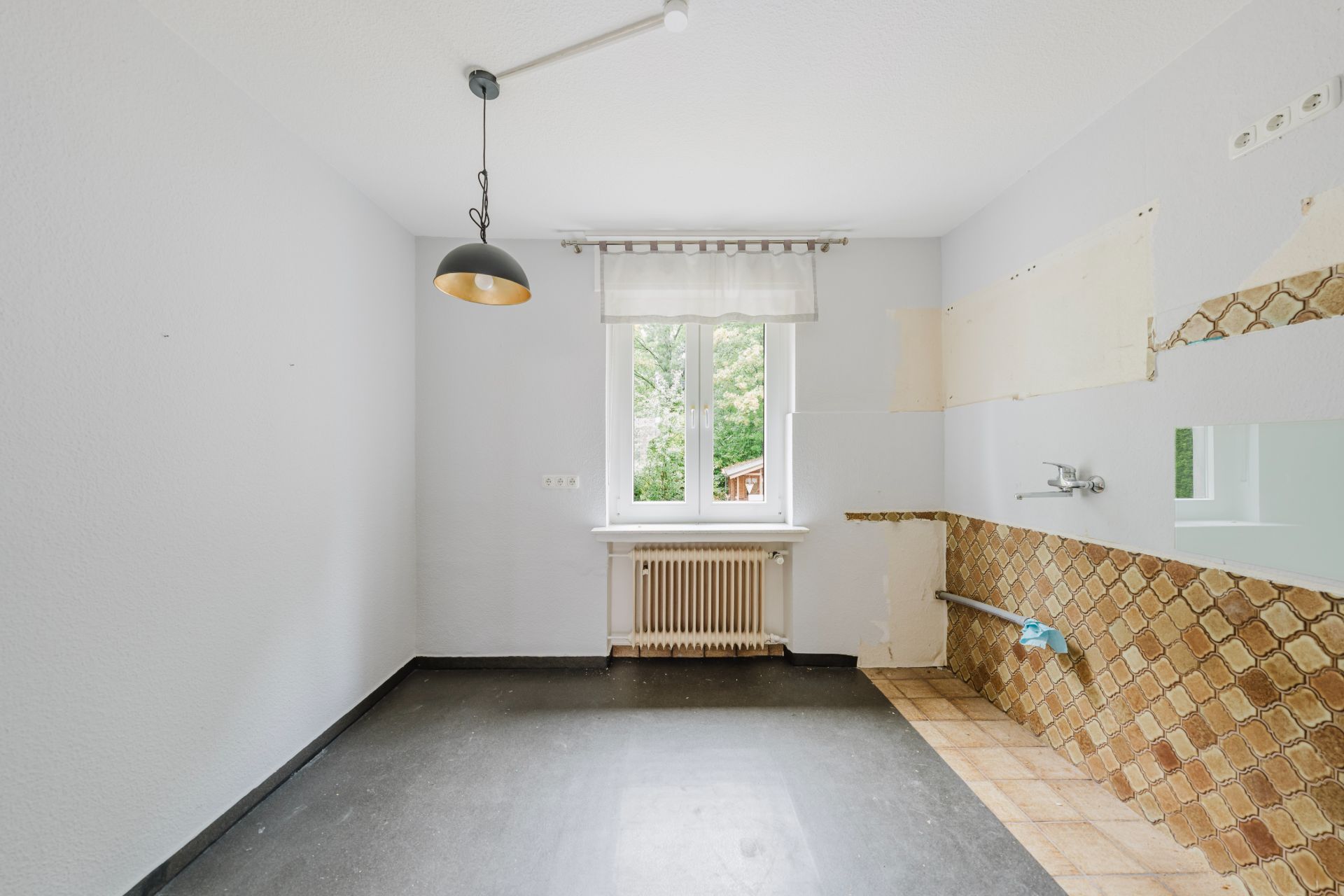 Leerer Raum mit Fenster, Heizkörper, Hängelampe und Fliesen an einer Wand ohne Kücheneinrichtung.