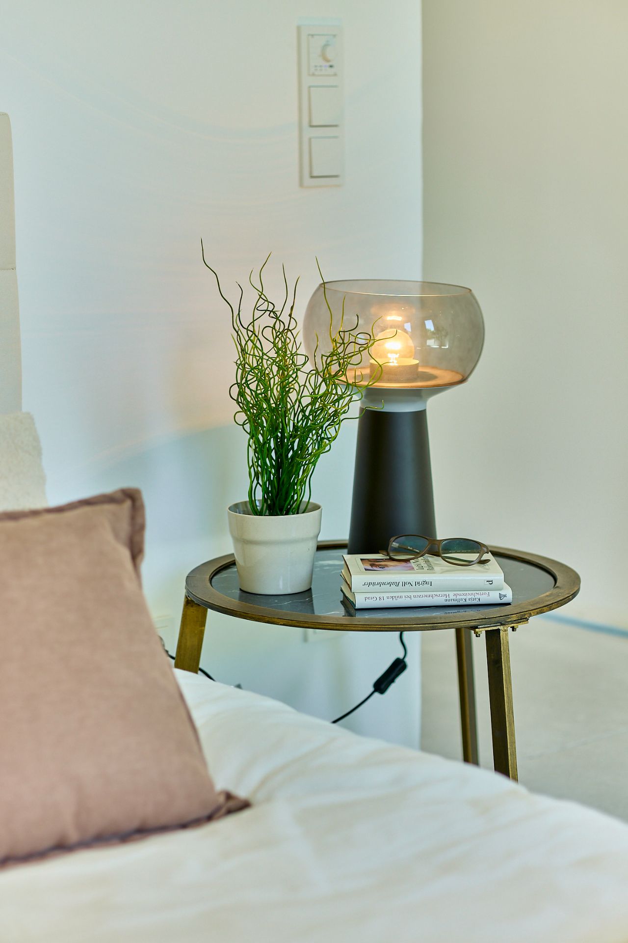Nachttisch mit Lampe, Pflanze, Büchern und Sonnenbrille neben einem Bett.
