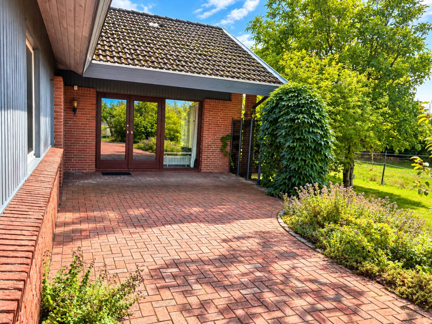 Gepflasterte Terrasse mit großen Glastüren, viel Grün und Büschen neben dem Haus.