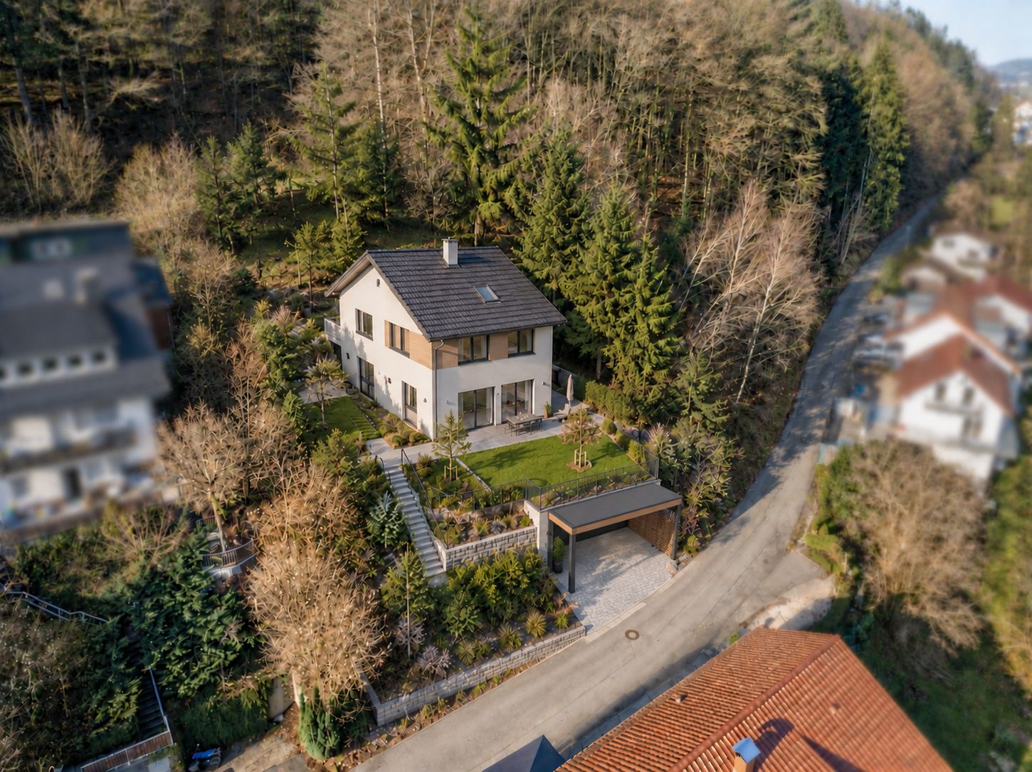 Luftaufnahme eines Einfamilienhauses mit Garten und Garage an einer Straße, umgeben von Bäumen.