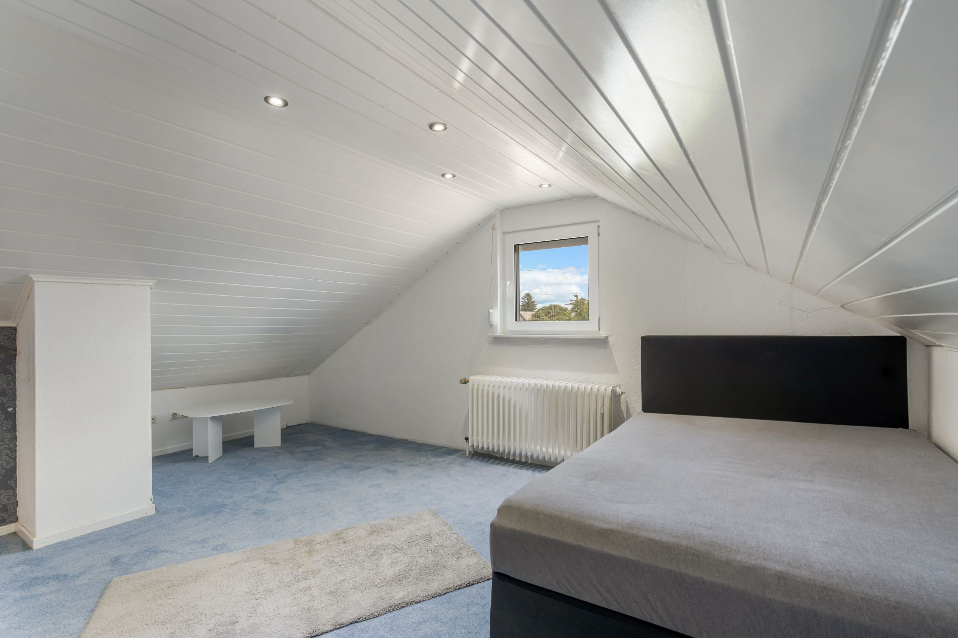 Helles Dachzimmer mit Bett, Teppich, Schreibtisch und Fenster, weiße Wände und Decke.