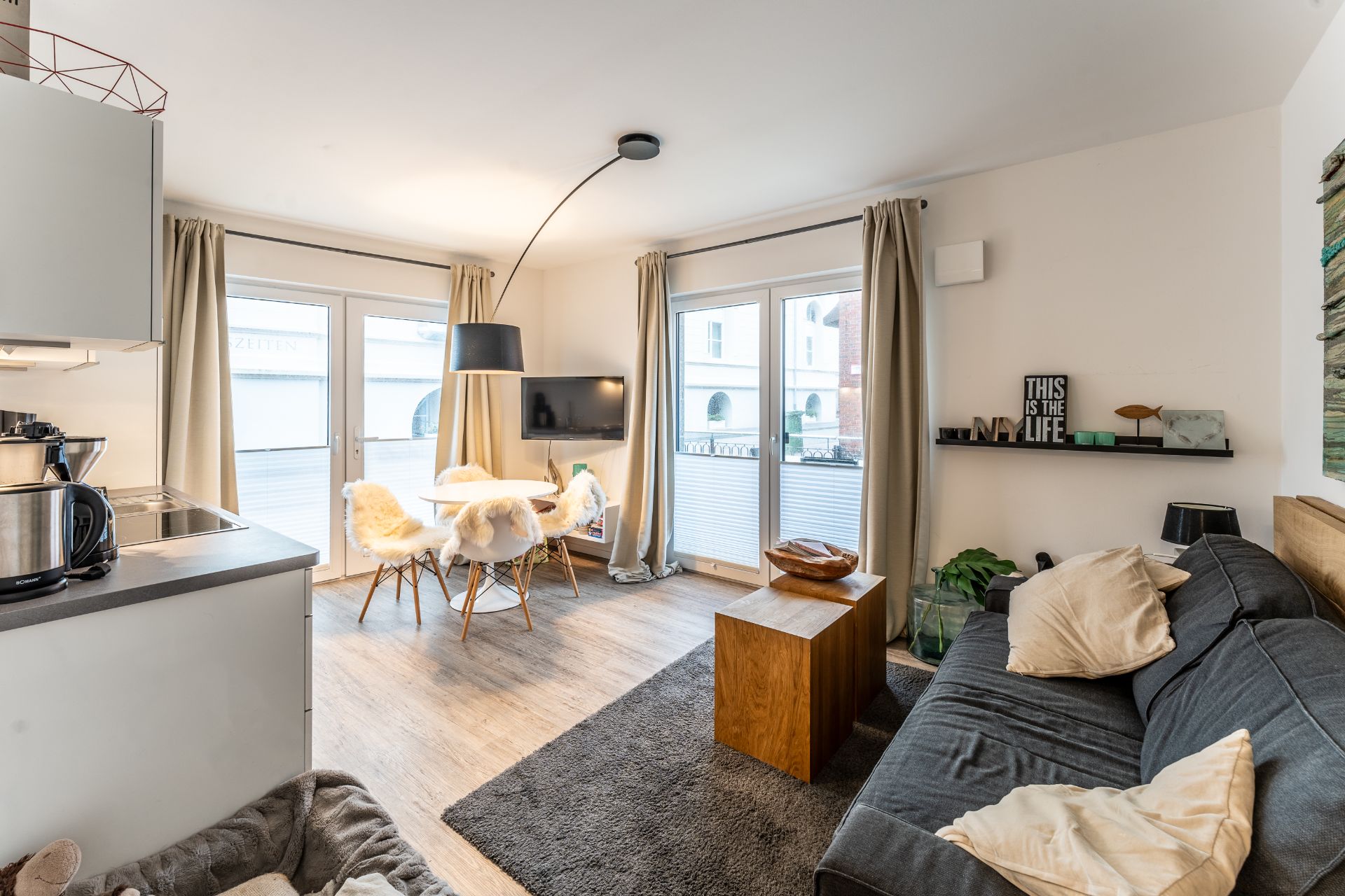 Modernes Wohnzimmer mit Sofa, Esstisch, Stühlen und großen Fenstern mit Vorhängen.