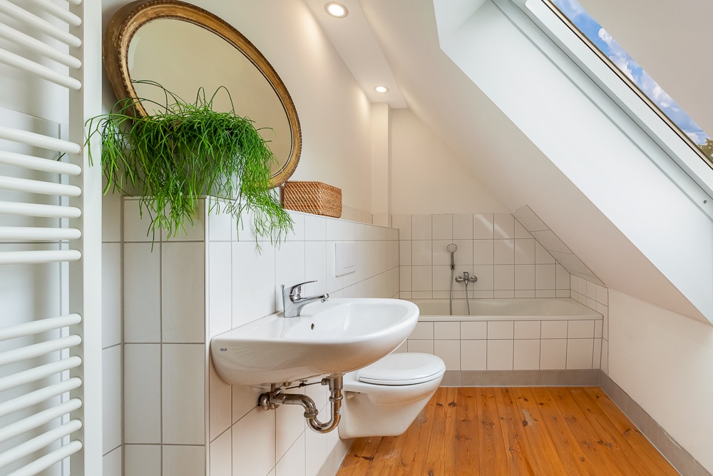 Modernes Badezimmer mit weißem Waschbecken, WC, Badewanne, Spiegel und Pflanze unter Dachschräge.