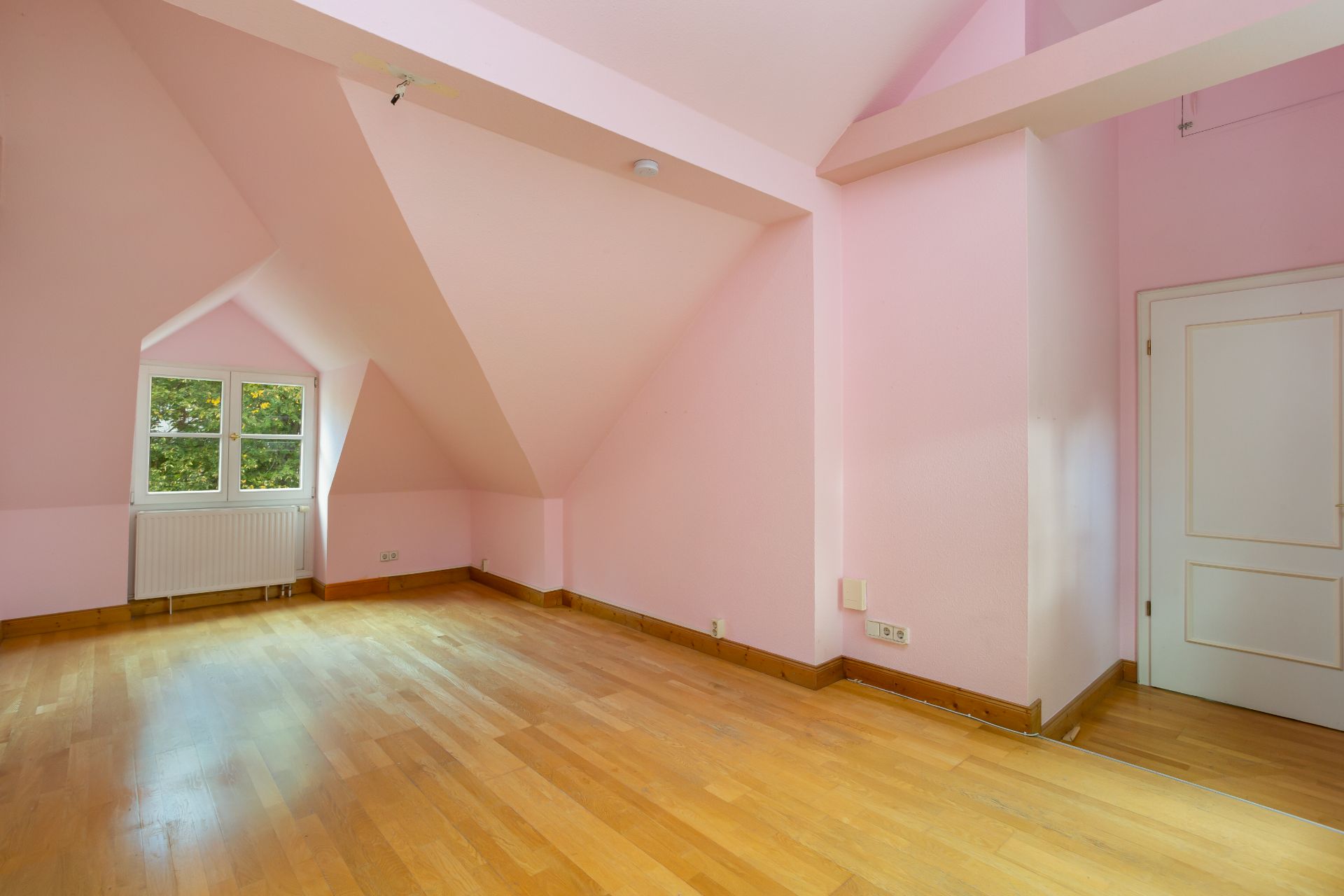Leeres Zimmer mit rosa Wänden, schrägen Decken und Holzfußboden, Fenster und Tür sichtbar.