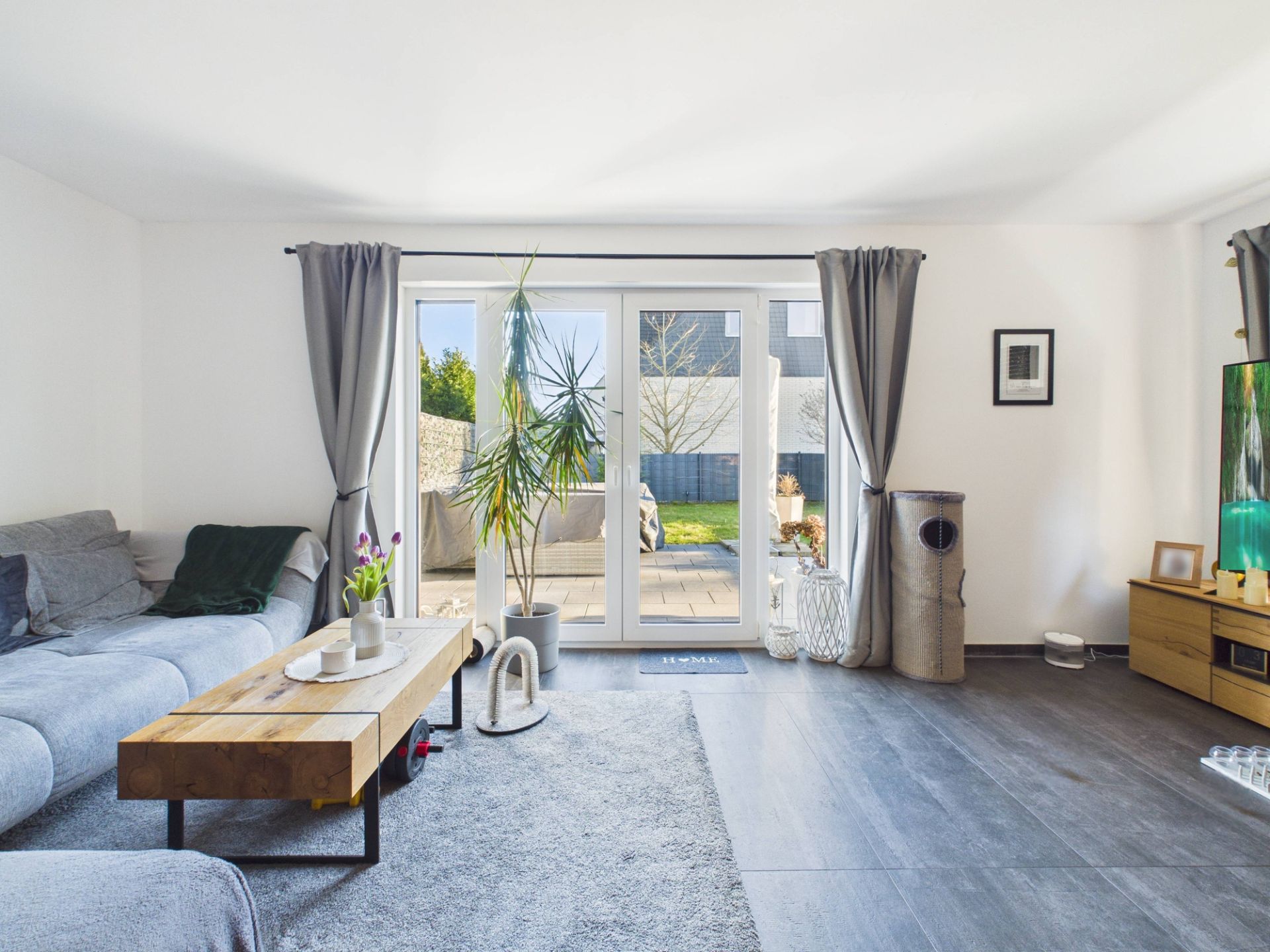 Helles Wohnzimmer mit grauem Sofa, Holztisch, großen Fenstern und Zugang zur Terrasse.