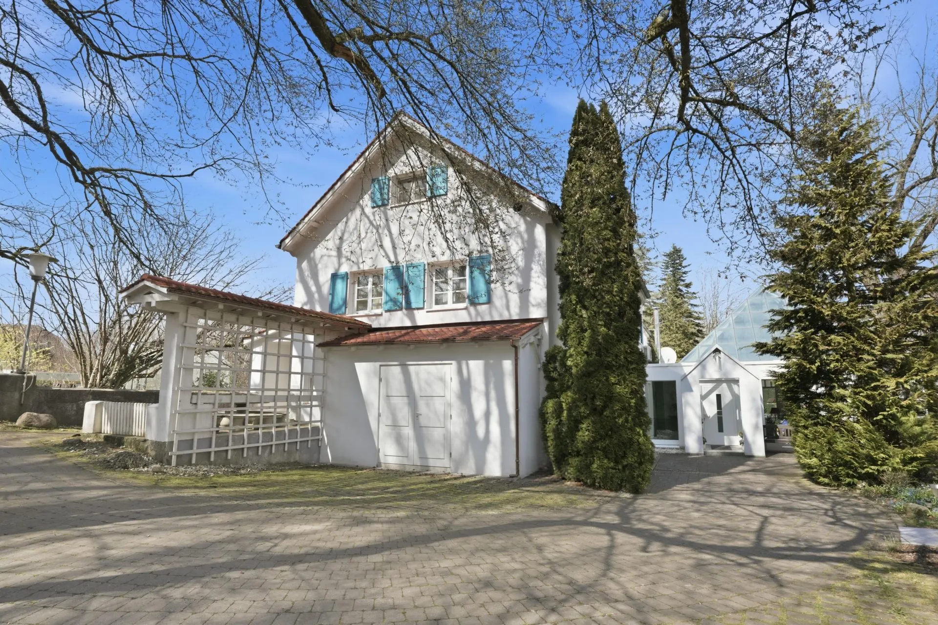 Weißes Wohnhaus mit blauen Fensterläden und Eingang, umgeben von Bäumen.