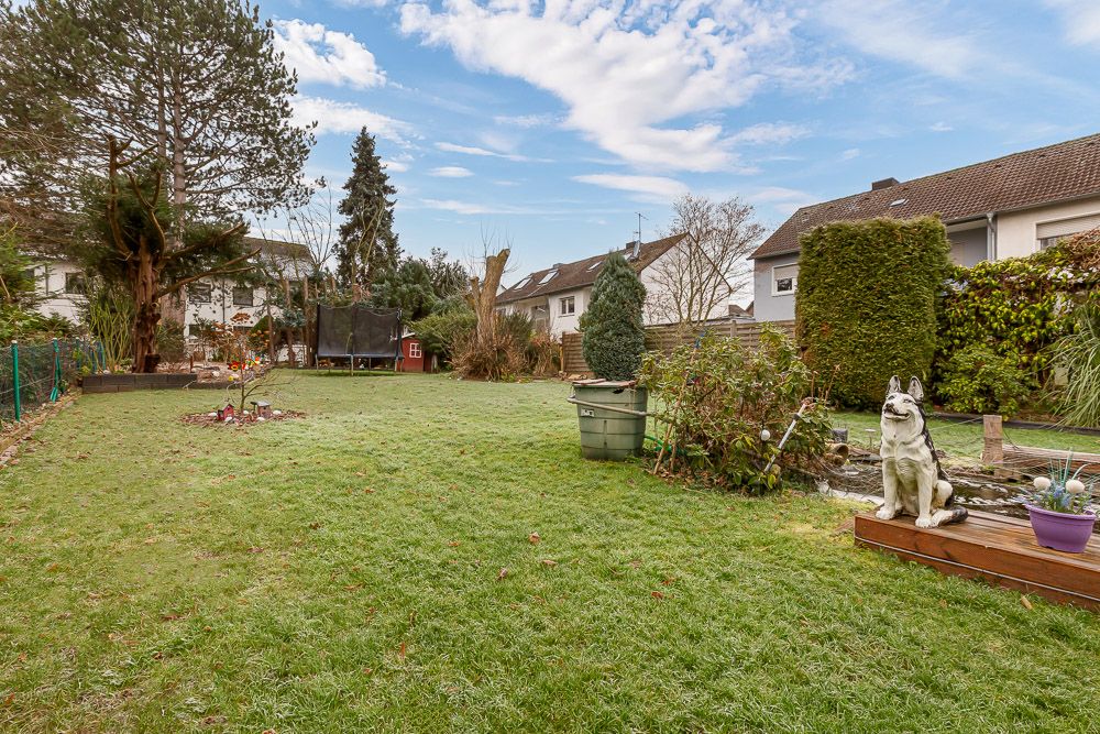 Großer Garten mit Rasen, Bäumen, Teich und Hundestatue vor Wohnhäusern.