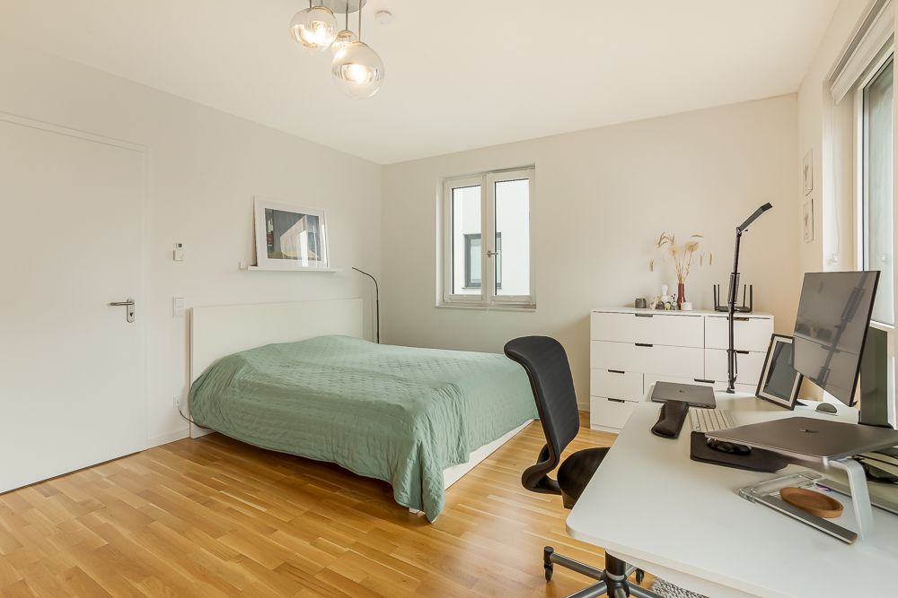 Helles Schlafzimmer mit grünem Bett, Schreibtisch, Stuhl und Kommode auf Holzfußboden.