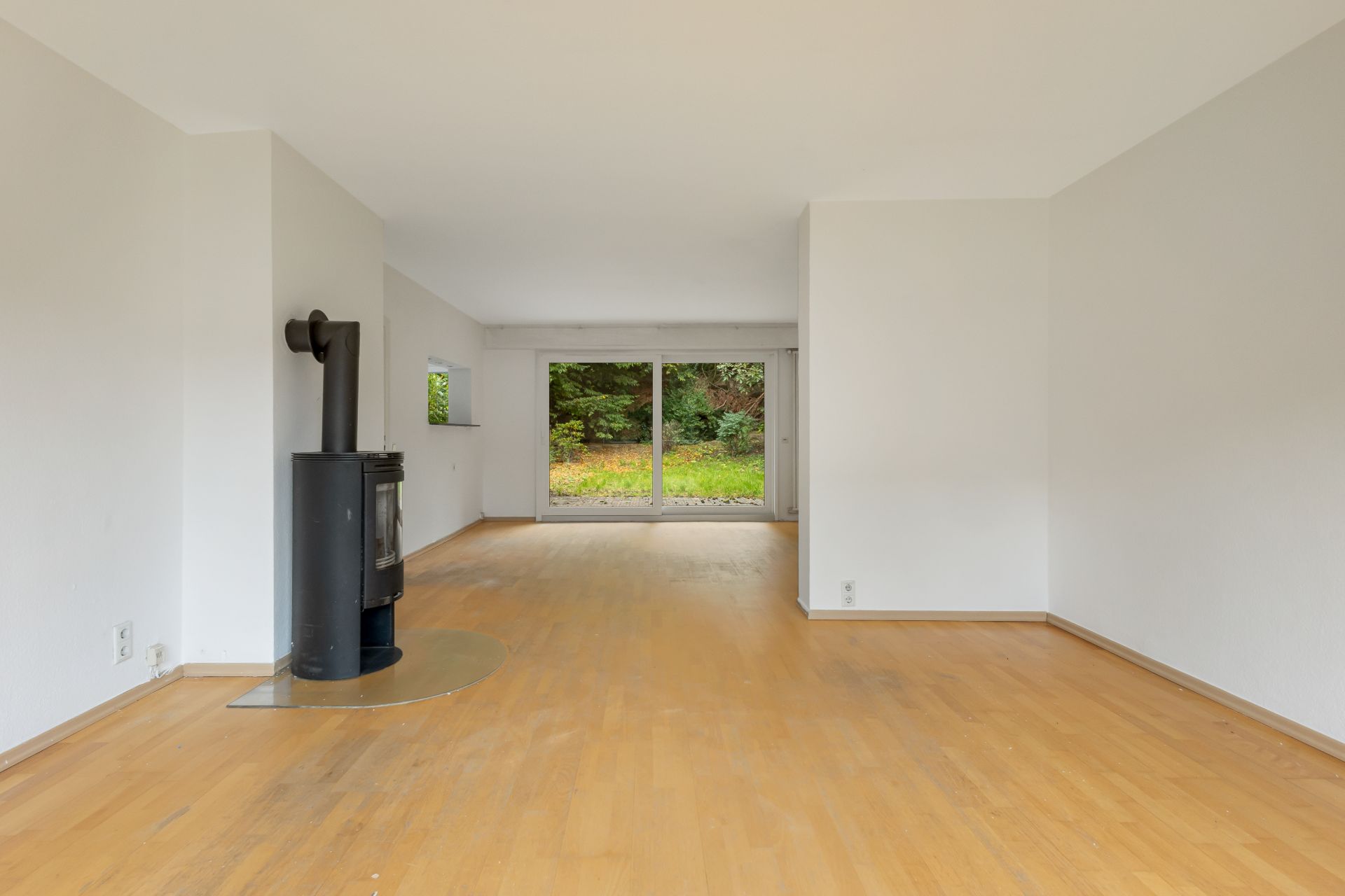 Leeres Wohnzimmer mit Holzboden, Kamin und großen Fenstern mit Blick ins Grüne.