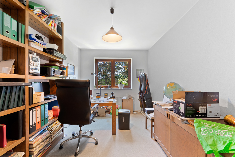 Arbeitszimmer mit Schreibtisch, Regalen, Stuhl und Fenstern, verschiedene Büroartikel und Bücher sichtbar.