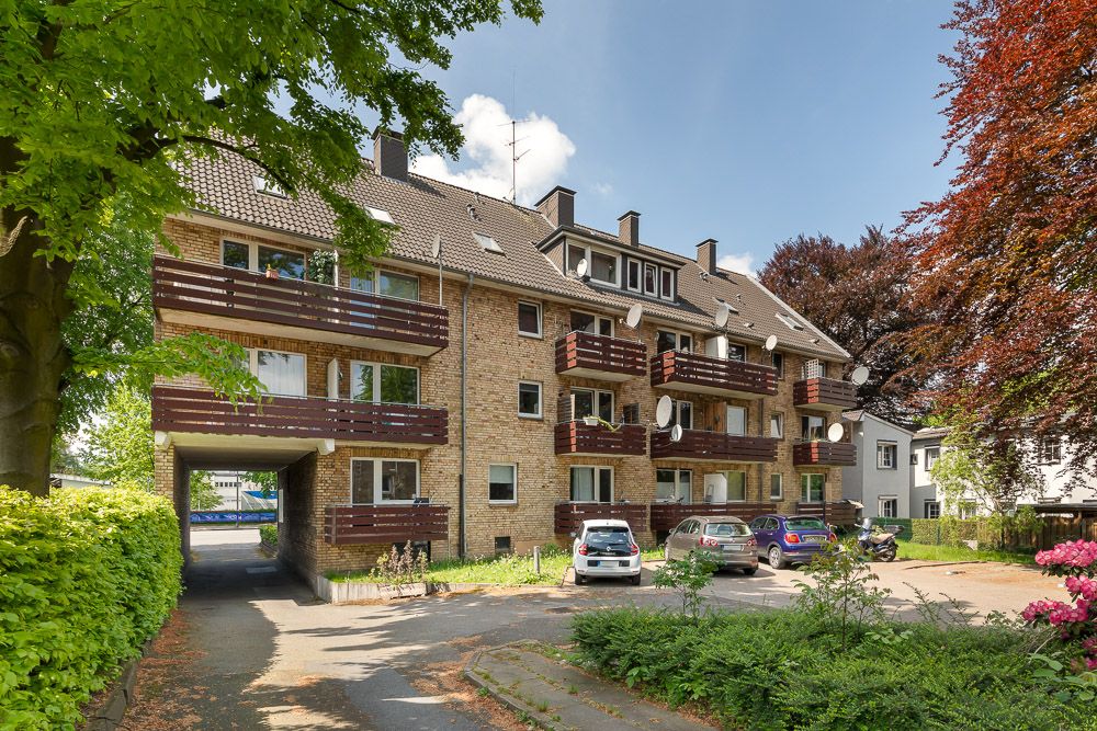 Mehrfamilienhaus mit Balkonen, geparkten Autos und Bäumen im Vordergrund.