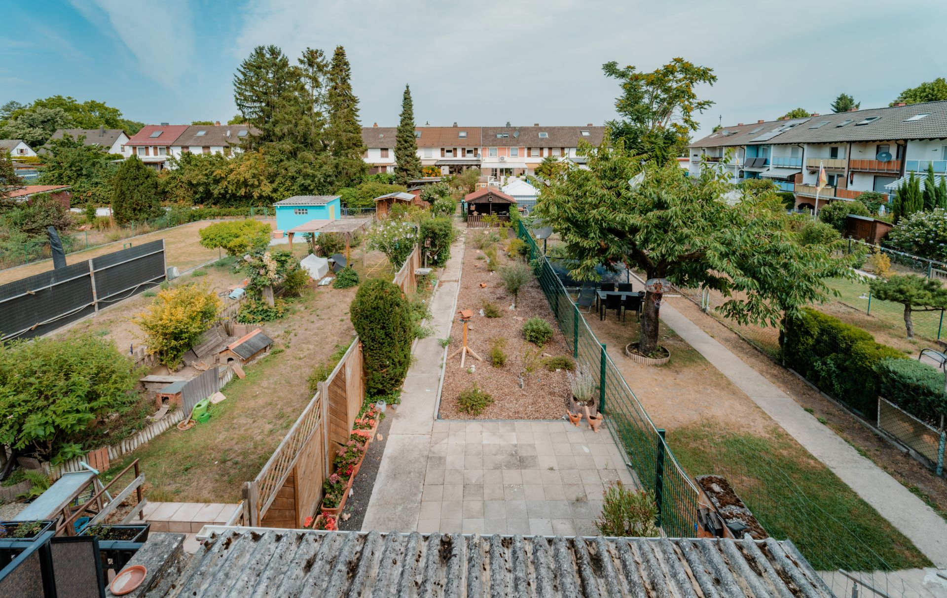 Blick auf einen langen Garten mit Wegen, Bäumen, Beeten und angrenzenden Reihenhäusern.
