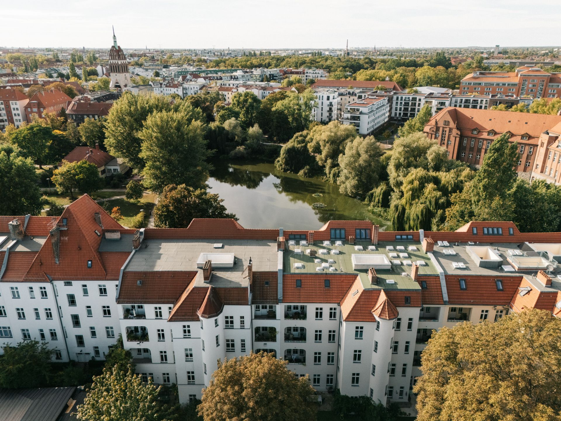 Weiße Wohnhäuser mit roten Dächern, dahinter ein Teich und viele Bäume.