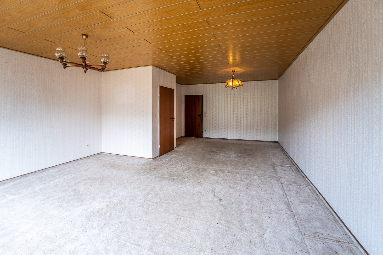 Leeres Zimmer mit Holzdecke, zwei Türen, Teppichboden und zwei Kronleuchtern.