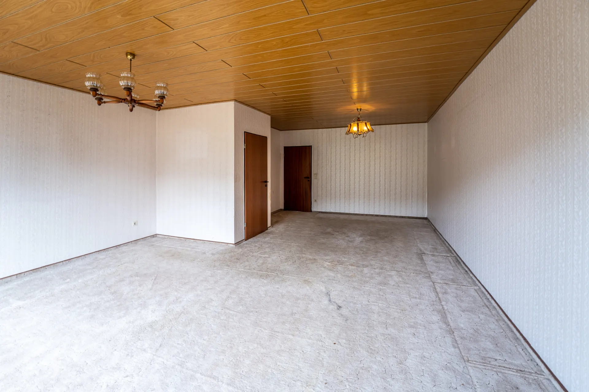 Leeres Zimmer mit Holzdecke, zwei Türen, Teppichboden und zwei Kronleuchtern.