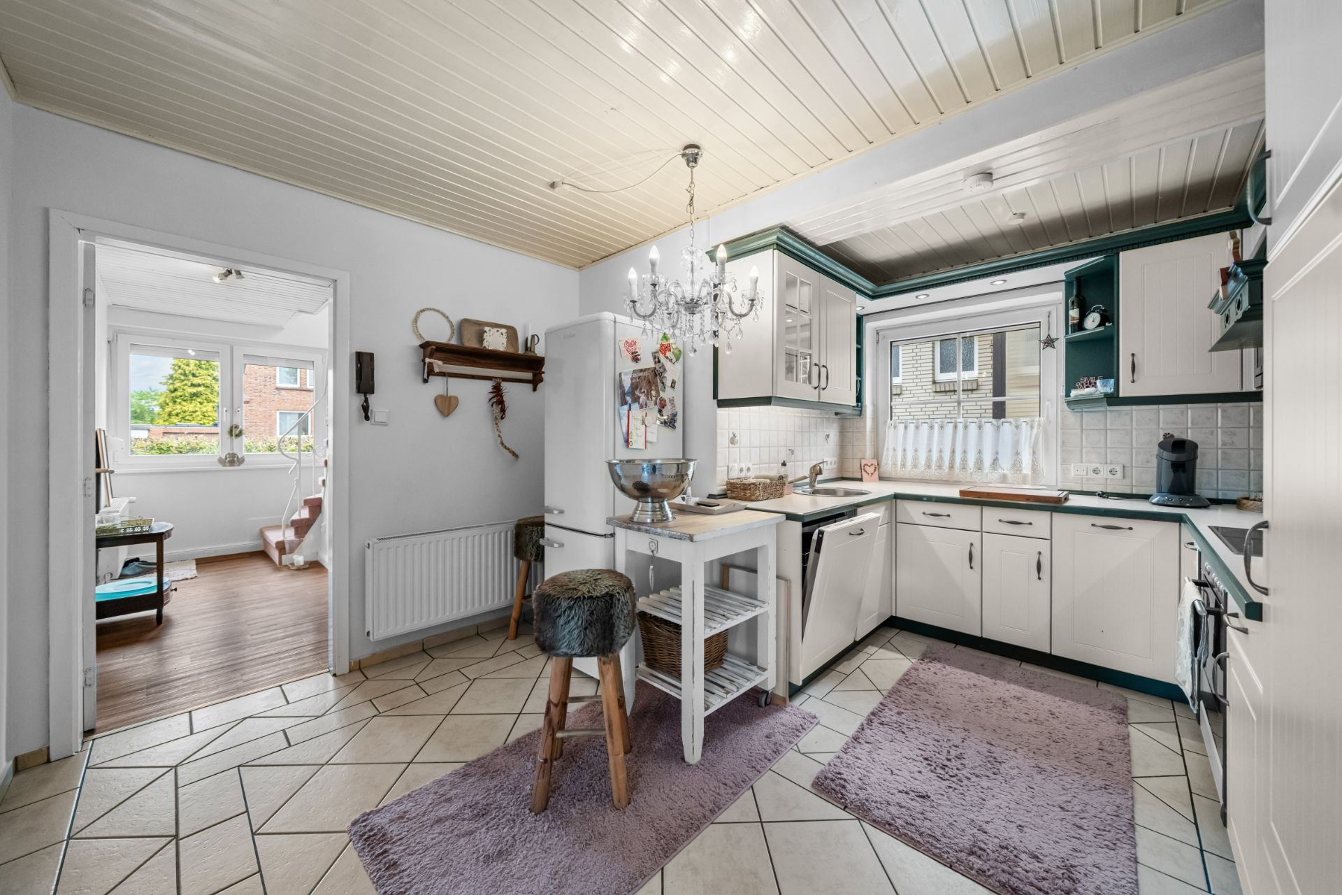 Helle Küche mit weißen Schränken, Kücheninsel, Hocker und Fenster, angrenzend an ein Wohnzimmer.